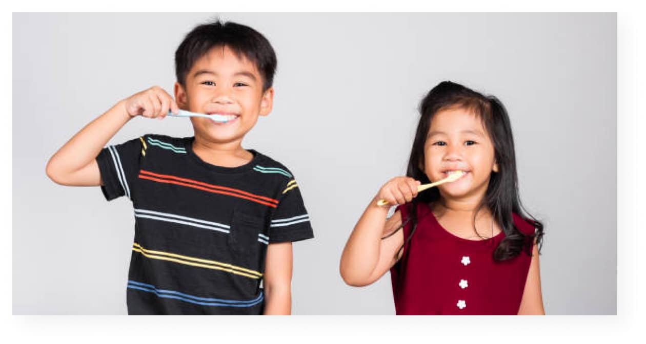 Two young children with brown hair, brushing their teeth in front of a plain gray background.